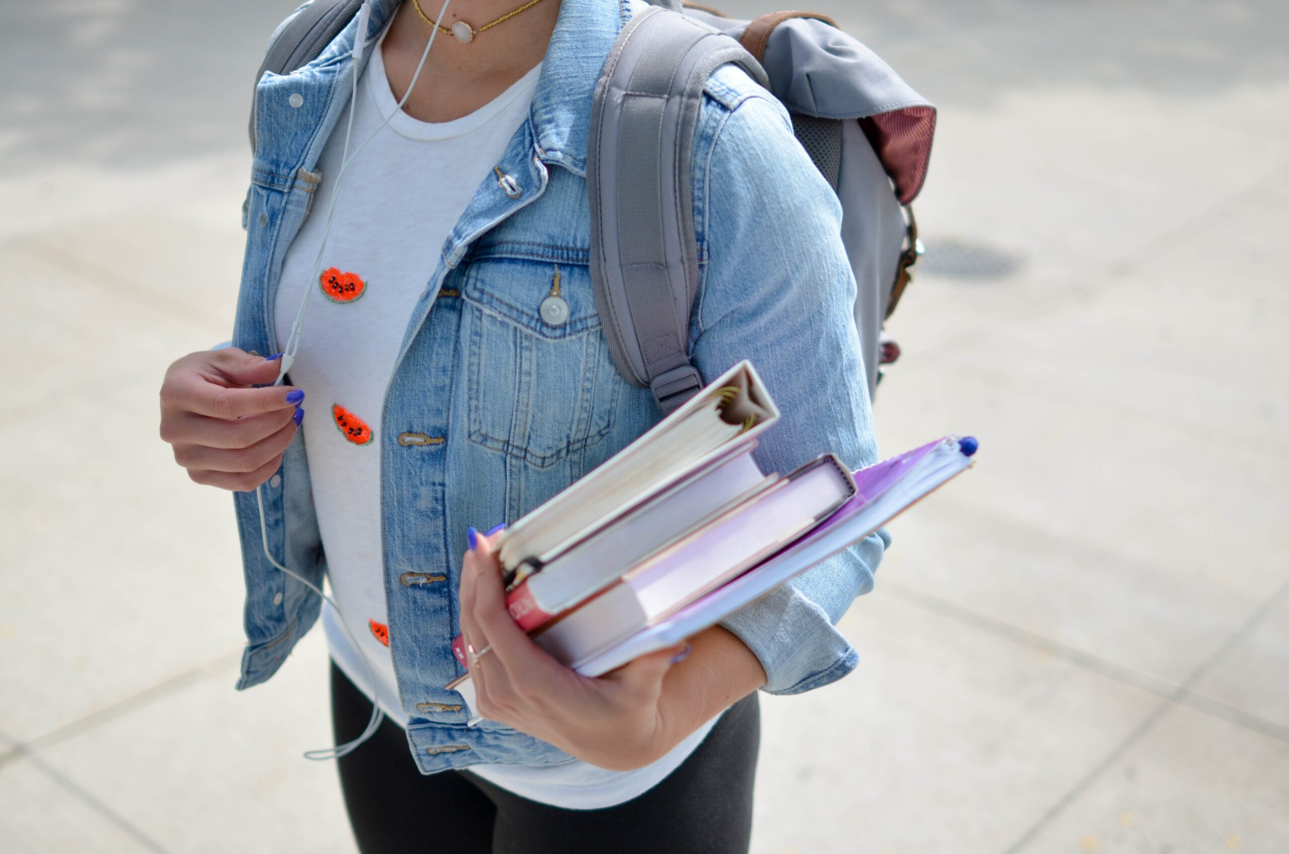 A young female high school student wearing a light blue jean jacket is walking to class wearing her backpack and carrying two notebooks and two books.
