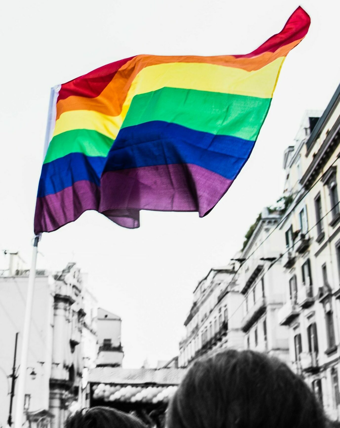 A pride flag waving above a crowd in a city street.